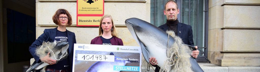 Three people, each holding three different objects. The woman on the left holds a small model of a porpoise caught in a net, the woman in the middle holds a sign with the petition, the man standing on the right holds a larger model of a porpoise caught in a net.