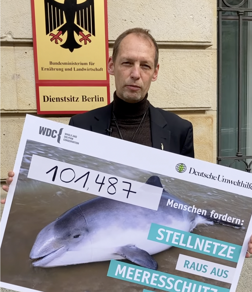 Ein Mann steht vor dem Bundesministerium für Ernährung und Landwirtschaft in Berlin und hält ein großes Schild mit den endgültigen Zahlen der Petition für die Rettung der Schweinswale. Die Zahl ist: 101487.