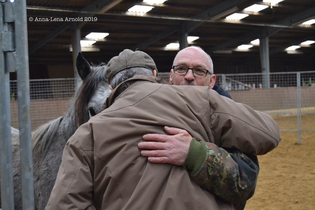 Two elderly men hug each other, with a gray horse in the background.