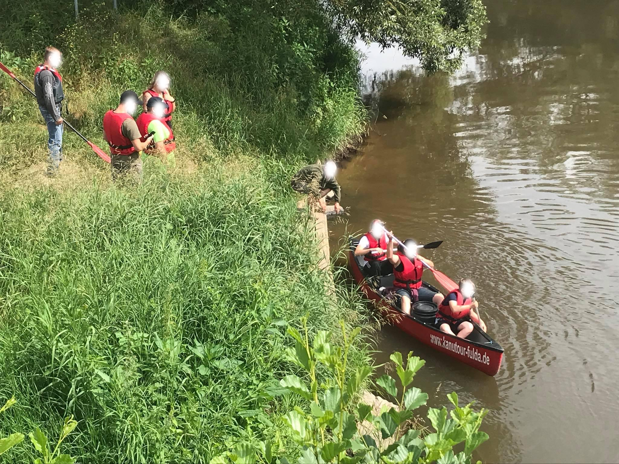 Ein rotes Kanu mit drei Personen schwimmt auf einem Fluss, während 5 weitere Personen am Ufer stehen und zusehen.