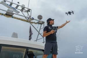 A man standing on the deck of a ship holds a remote control and flies with a drone flying above him. He reaches out his hand to the drone. The sky is cloudy.