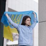 Eine nach vorne blickende, junge Frau mit braunem Haar und hellblauem Pullover hält mit erhobenen Armen eine Ukraine Flagge über und hinter ihren Kopf.