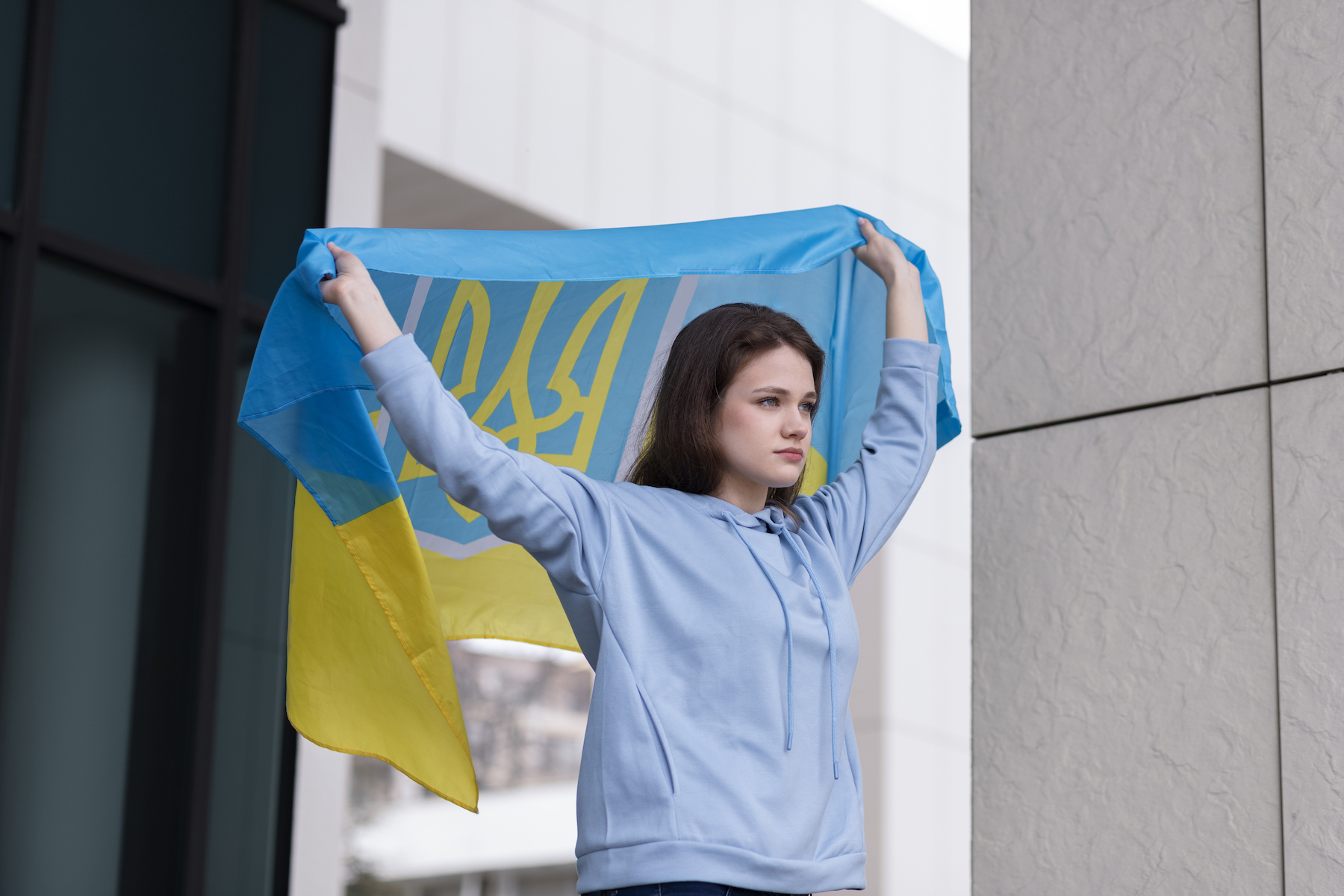 A looking forward young woman with brown hair and light blue sweater is holding a Ukraine flag above and behind her head with raised arms.