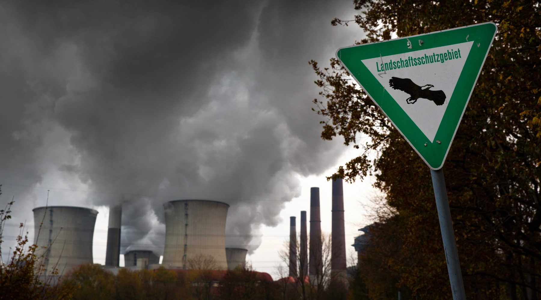 Im Vordergrund steht ein Schild mit der Aufschrift "Landschutzgebiet". Im Hintergrund sieht man ein Kraftwerk, welches Rauch ausstößt.