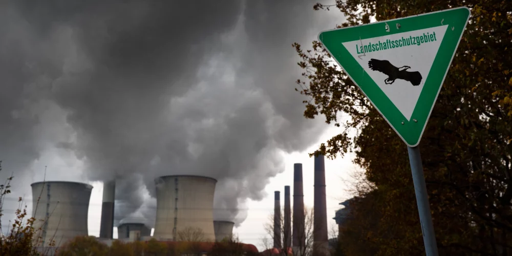 Im Vordergrund steht ein Schild mit der Aufschrift "Landschutzgebiet". Im Hintergrund sieht man ein Kraftwerk, welches Rauch ausstößt.
