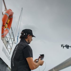 A man standing on a boats deck with a remote control and a drone is flying above him.